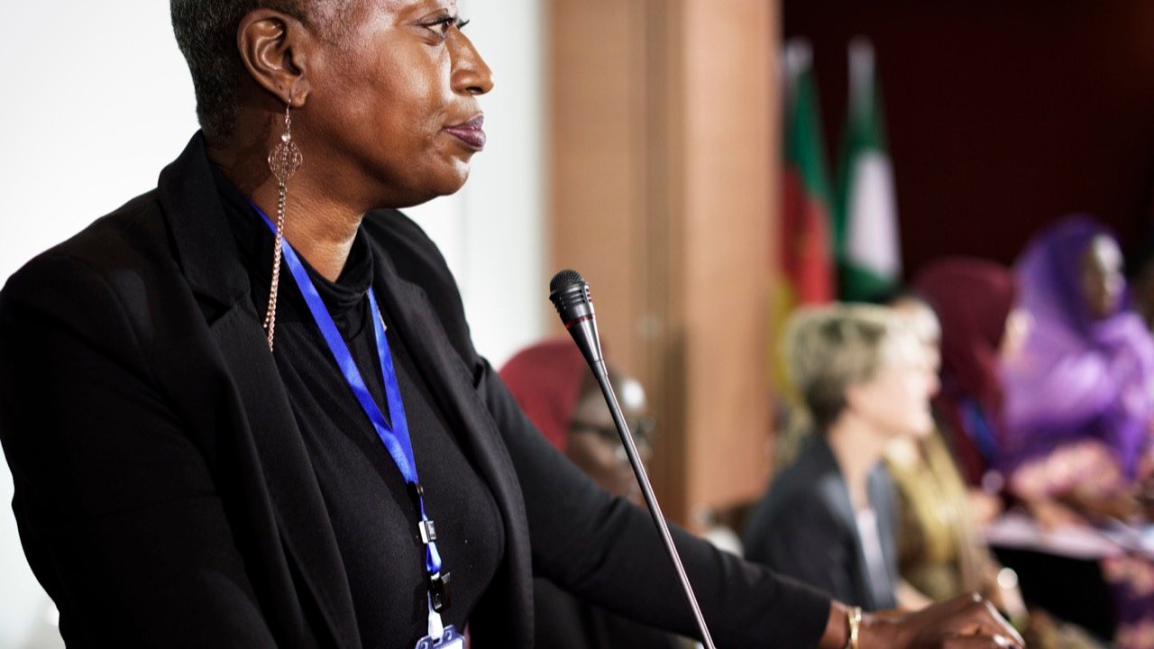 Une femme parle au micro lors d'une conférence, des drapeaux en arrière-plan